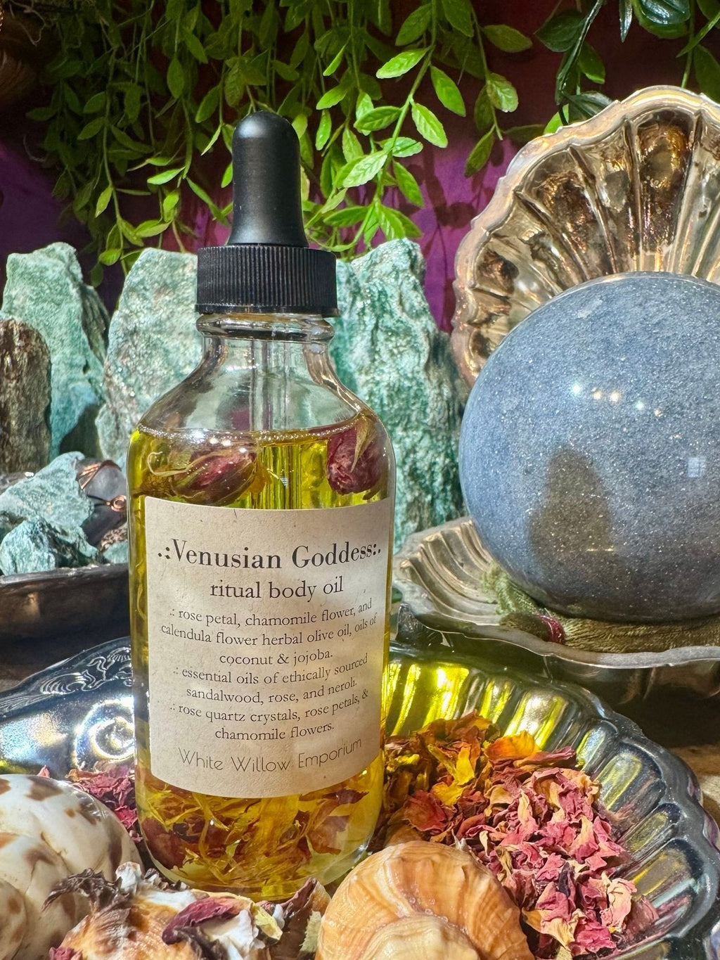 A bottle of 'Venusian Goddess' ritual body oil on a table with crystals, shells, and a crystal ball.
